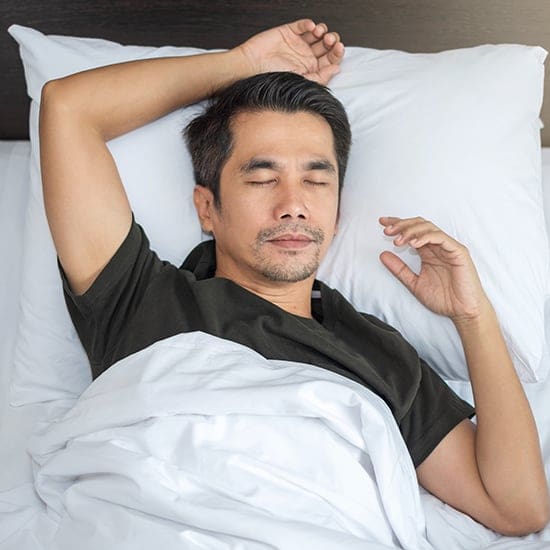 A man with short dark hair and facial hair is sleeping peacefully on a bed. He is lying on his back, with one arm resting behind his head and the other placed gently on his chest. He is covered by a white blanket and wears a dark-colored shirt.