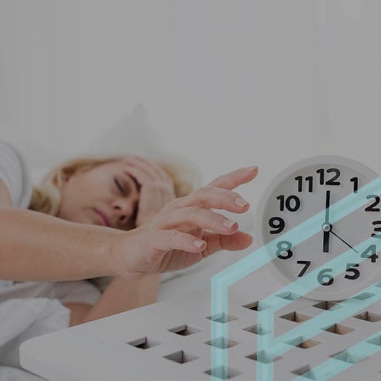 A person lying in bed reaches out with a hand toward a white analog alarm clock displaying a time of 7:50. The person appears to be trying to turn off or silence the alarm. Their face shows a tired expression, and they rest their head on the other hand.
