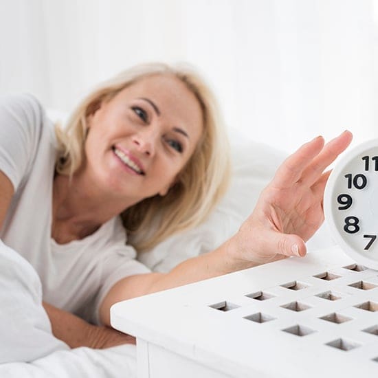 A smiling woman with blonde hair lies in bed, reaching out to turn off an alarm clock on a nightstand. She is wearing a white shirt and the room has a bright, airy atmosphere.
