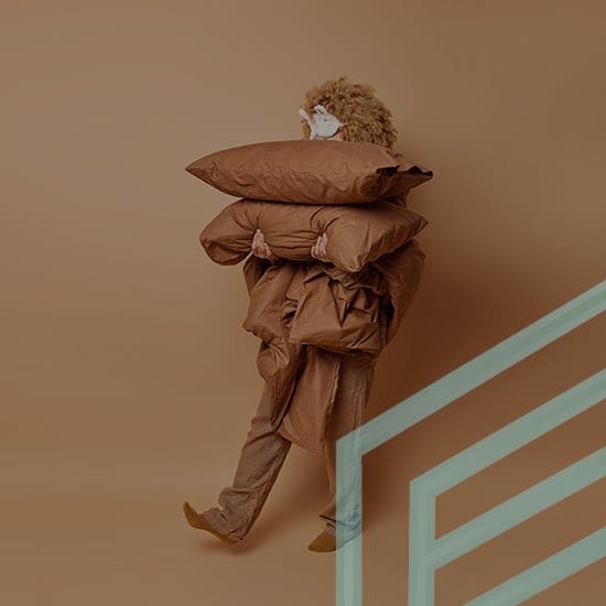 A person with curly hair, wearing brown clothing and shoes, carries two large brown pillows. The background is a solid brown, and part of a light green geometric design appears in the foreground on the right side.