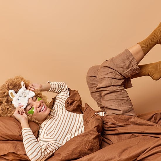 A person with curly hair is reclining on a brown bed, wearing striped pajamas, a plush unicorn sleep mask, and under-eye patches. Their legs are playfully raised, and they have brown socks on, set against a neutral background.