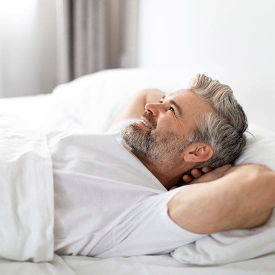 A bearded man with gray hair lies in bed with a relaxed expression, his arms resting behind his head. He is wearing a white shirt and looking towards the window. The room has a bright and airy feel with white bedding and curtains.