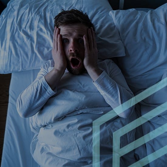 A man lying in bed with white sheets and pillows, wearing a white long-sleeve shirt. He appears to be in shock, with his mouth open and hands on his cheeks. The background is dimly lit, adding to the surprised or startled expression on his face.