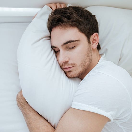 A man with short dark hair and a beard sleeps peacefully, hugging a white pillow and lying on white bedsheets. He is wearing a white T-shirt and is positioned on his side, with his eyes closed and face relaxed.