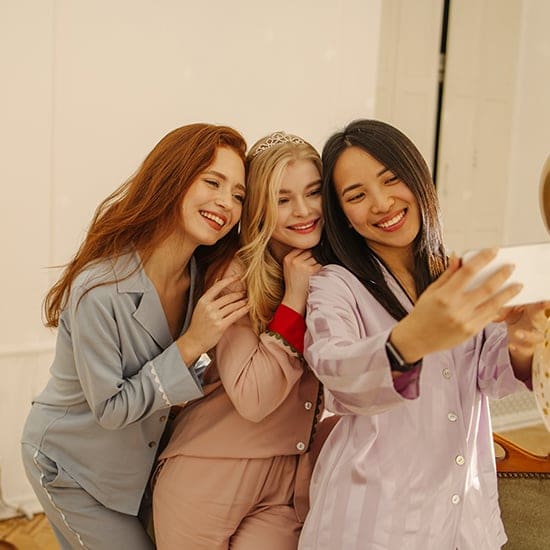 Three smiling women in pajamas take a selfie together. One woman with red hair is in blue pajamas, the middle woman with blonde hair wears a crown and pink pajamas, and the woman on the right with black hair is in lavender pajamas. They appear to be enjoying a fun moment.