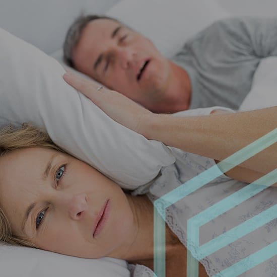 A woman lies awake in bed, looking frustrated while a man next to her sleeps with his mouth open. The photo suggests the woman is disturbed by the man's snoring. The scene is set in a bedroom, both individuals lying on white pillows.