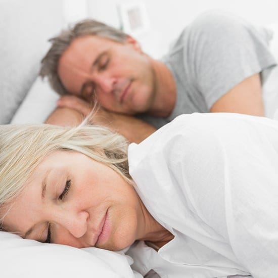 A middle-aged couple with gray hair is peacefully sleeping in bed. They are lying on their sides, with the woman in the foreground and the man behind her. Both are dressed in light-colored sleepwear, and the setting appears relaxed and comfortable.