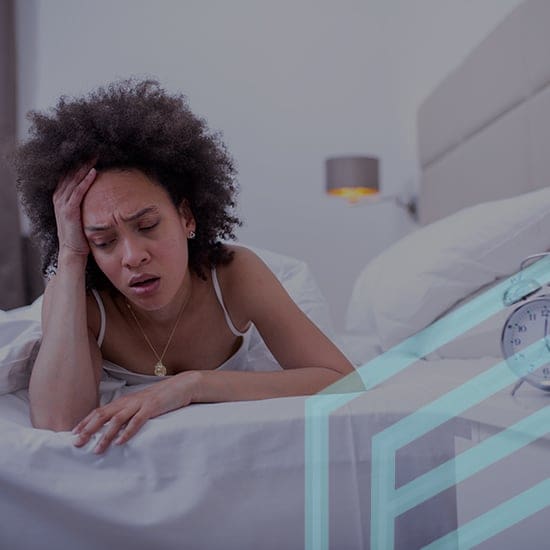 A woman sits on her bed, leaning forward with her hand on her forehead, appearing distressed or in pain. An alarm clock is visible on the bedside table, and a lamp is turned on in the background. The scene is in a bedroom with white bedding and neutral tones.