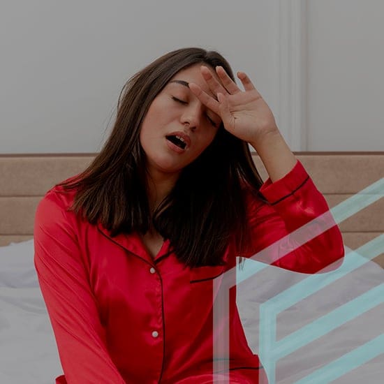 A woman in pink satin pajamas sits on a bed, looking tired and yawning. Her right hand is raised to her forehead as she appears to be rubbing it. The background shows a white wall and a beige headboard. The atmosphere suggests she has just woken up.
