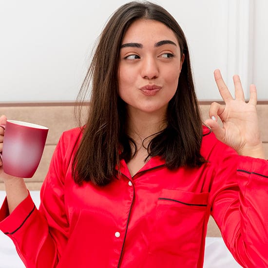 A woman wearing a red satin pajama top is holding a red mug with a white interior in one hand and making an "OK" gesture with the other. She has long dark hair and appears to be standing in a bedroom, giving a playful expression.