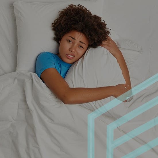 A person with curly hair, wearing a blue shirt, lies in bed hugging a pillow with a distressed expression. The bed has white sheets and pillows. A faint geometric pattern is visible in the lower right corner of the image.