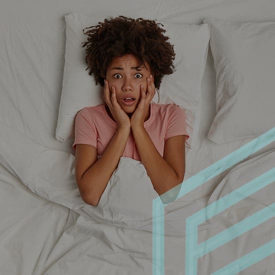 A young person with curly hair looks surprised and concerned while lying in bed, hands pressed to the sides of their face. They are wearing a pink shirt and are partially covered by a white blanket. The background shows white pillows and bedding.