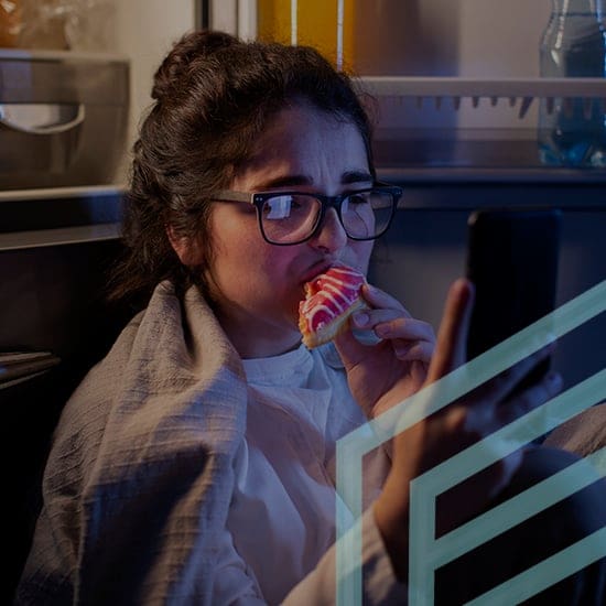 A woman with glasses, wrapped in a blanket, sits in front of an open refrigerator at night. She is holding a smartphone with one hand and eating a doughnut with red icing in the other. The refrigerator light illuminates her face and surroundings.