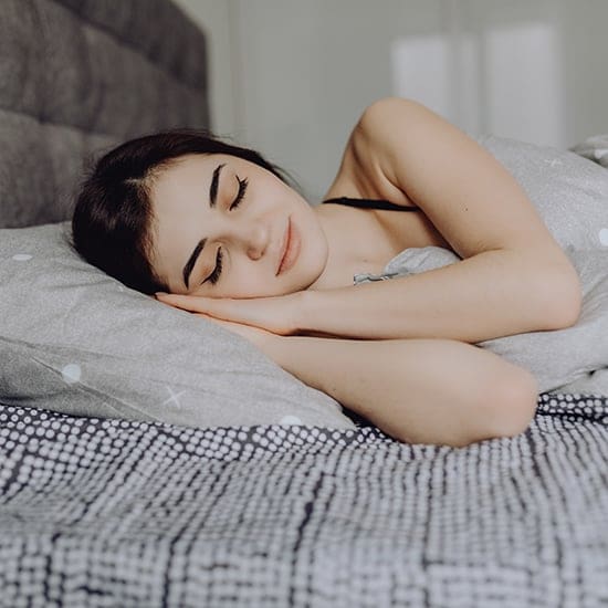 A person with dark hair is peacefully sleeping on their side in a bed, covered with a gray and white polka-dotted blanket. Their head rests on a pillow with a matching pattern, and their hands are gently tucked under the pillow. The room appears softly lit.