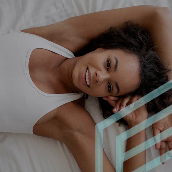 A woman with curly hair lies on her back on a white bed, wearing a white tank top. She is smiling and facing the camera with one arm raised above her head. The image has a relaxed and cheerful atmosphere.