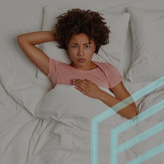 A woman with curly hair lies in bed on her back with a concerned expression. She wears a pink shirt and has one hand on her chest and the other behind her head. White pillows and sheets surround her. A geometric, translucent design overlays part of the image.