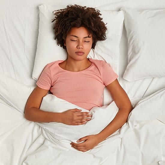 A woman with curly hair lies in bed on her back, eyes closed, with one hand resting on her stomach and the other under a white blanket. She wears a light pink t-shirt, and the bedding is all white. The setting appears serene and comfortable.