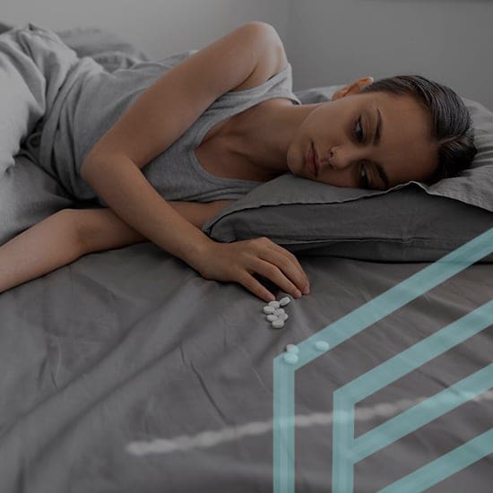 A person is lying on a bed, appearing somber and reflective. They are resting their head on a pillow and looking at small white pills on the bed beside them. The room has a minimalist design with gray bedding.