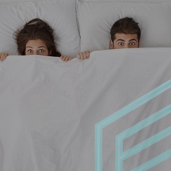A man and a woman lie in bed side by side, peeking out from under a white blanket with wide eyes. They are covered up to their noses. The blanket has a faint turquoise geometric outline on the right side.