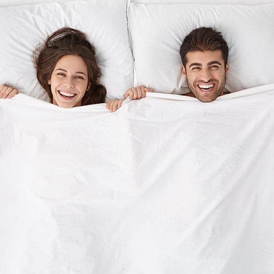 A woman and a man are lying in bed, smiling broadly while covered up to their chests with a white bedsheet. The background is a plain white wall. Both have dark hair and appear happy and relaxed.