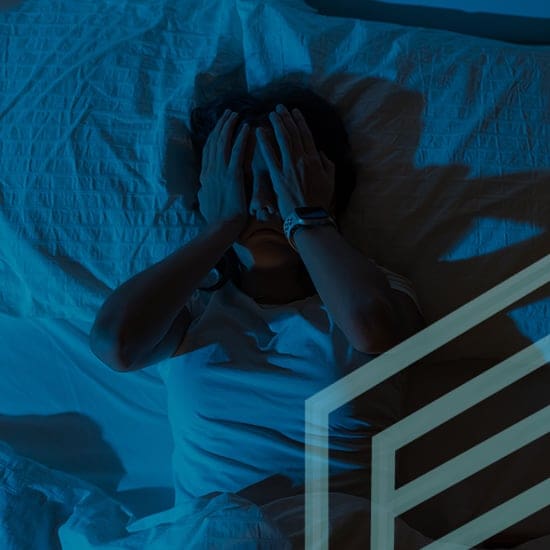 A person lying in bed at night covers their face with their hands in distress. The scene is dimly lit with a blue hue, and a geometric shadow falls across the lower part of the image. The bed has a textured white sheet and pillow.