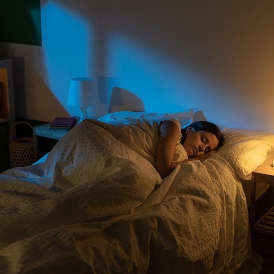 A woman sleeps peacefully on her side in a bed with white sheets and pillows. The room is dimly lit with a warm light from a bedside lamp on a small table, and a soft blue light creates a calm and serene atmosphere. A basket and some books are in the background.