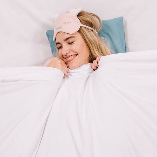 A woman with closed eyes and a joyful smile is lying in bed under a white blanket. She is wearing a pink sleep mask with a cute design and her head is resting on a teal pillow. She appears to be in a state of relaxation and comfort.