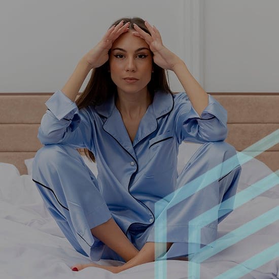 A woman with long dark hair sits cross-legged on a bed, wearing blue satin pajamas with black piping. She rests her hands on her forehead and looks directly at the camera. The background shows a beige headboard and white walls. Geometric lines overlay the image.