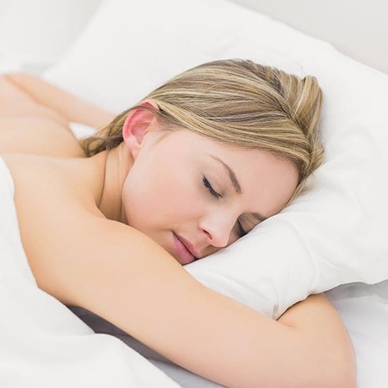 A person with light hair is sleeping peacefully on their side with eyes closed. They are resting their head on a white pillow and covered with a white blanket, suggesting a comfortable and restful sleep environment.