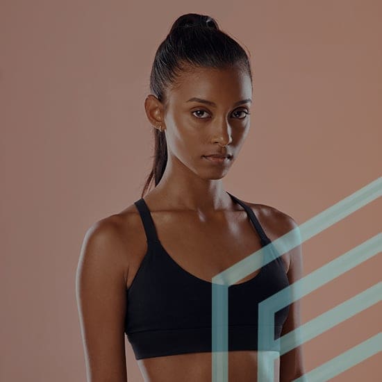 A woman with long, dark hair tied in a high ponytail, wearing a black sports bra, stands against a light brown background. She looks directly into the camera with a neutral expression, and a geometric design with light blue lines overlays the bottom right corner.