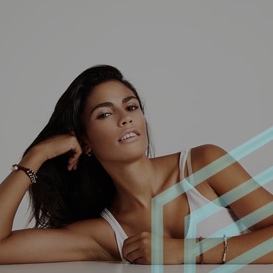 A woman with long dark hair, wearing a white sleeveless top, is leaning on a surface with her left arm propped up and her right arm relaxed. She is looking directly at the camera and smiling slightly. The background is plain gray with a blue geometric overlay in the lower right corner.