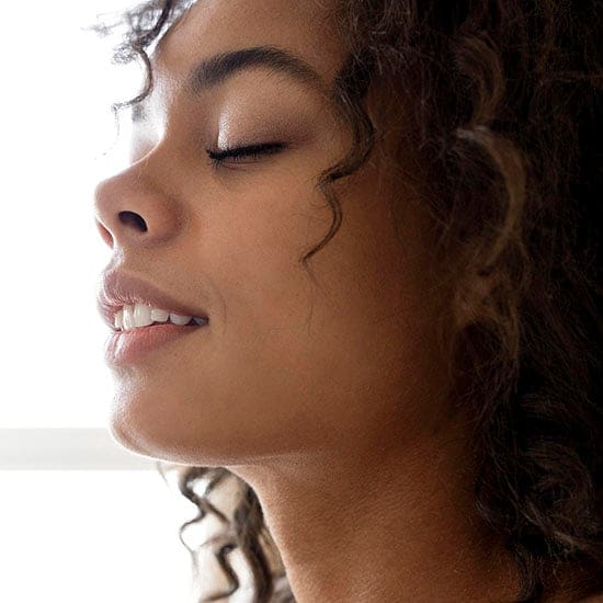 A close-up image of a person with curly hair and closed eyes, facing left. They have an expression of contentment or serenity. The background is softly lit, emphasizing the calm and peaceful mood of the scene.