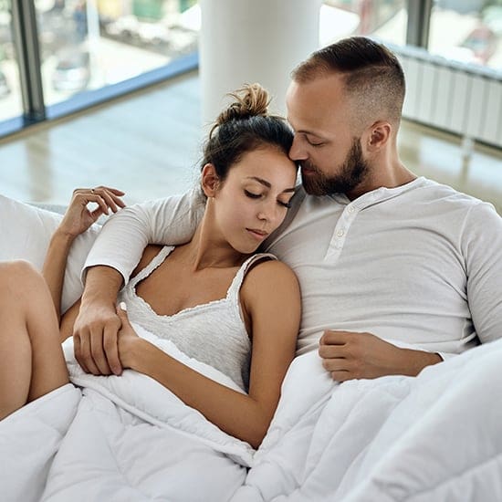 A couple in white clothing is cuddling on a bed covered with a white comforter. The woman has her eyes closed, resting her head on the man's chest, while the man holds her gently, with his head resting on hers. They are in a bright room with large windows.