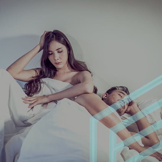 A woman and a man are lying in bed under white blankets. The woman is sitting up, looking thoughtful with her hand on her head, while the man is lying on his back, sleeping peacefully. The image is soft lit and conveys a mood of contemplation.