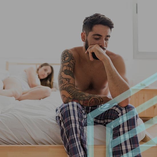 A shirtless man with tattoos and wearing plaid pants sits on the edge of a bed looking thoughtful. A woman lies on the bed in the background, partially covered by a blanket, gazing at him. The bedroom is simple with wooden furniture and natural light.