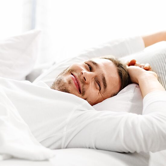 A person with a light beard is lying in bed, smiling while looking at the camera. They are wearing a white shirt, resting their head on a pillow in a bright room, embodying the tranquility often sought through sleep therapy. Their hands are behind their head, suggesting a relaxed and comfortable posture. A person with a light beard is lying in bed, smiling while looking at the camera. They are wearing a white shirt, resting their head on a pillow in a bright room, embodying the tranquility often sought through sleep therapy. Their hands are behind their head, suggesting a relaxed and comfortable posture.