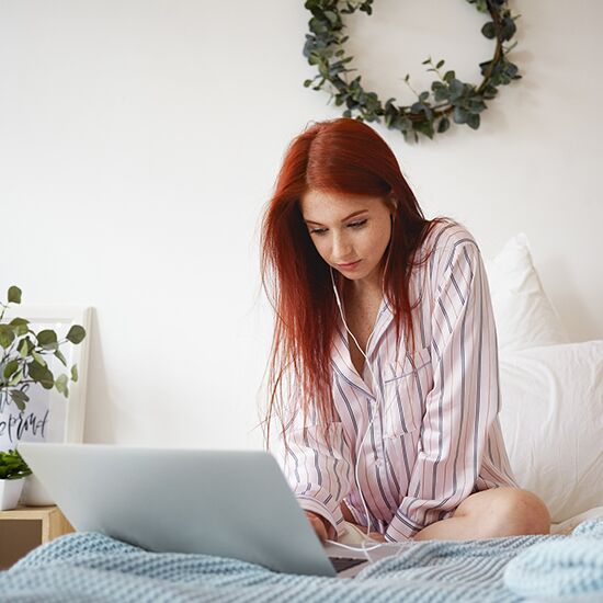 A person with long red hair wearing striped pajamas sits on a bed, using a laptop for hypnotherapy. They have earphones on, perhaps seeking relief from insomnia. The bed features a blue knitted blanket, and a leafy wreath hangs on the white wall beside a nearby plant. A person with long red hair wearing striped pajamas sits on a bed, using a laptop for hypnotherapy. They have earphones on, perhaps seeking relief from insomnia. The bed features a blue knitted blanket, and a leafy wreath hangs on the white wall beside a nearby plant.