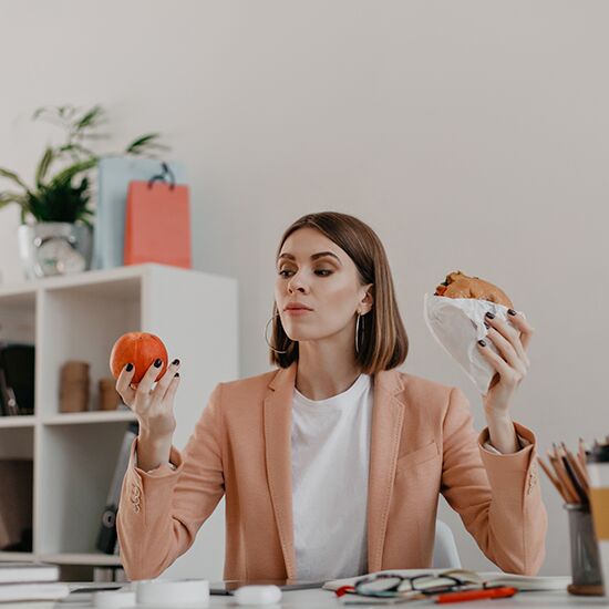 Female manager posing in white office. Lady looks at apple reluctantly, wanting to eat burger.