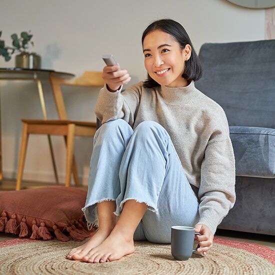 Portrait of beautiful asian girl sitting at her home and watching tv, holding remote, smiling and laughing, feeling comfort and warmth at her apartment
