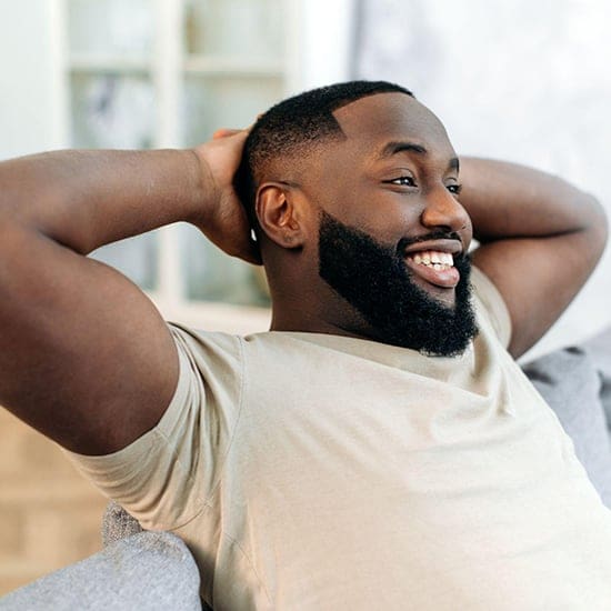 A smiling man with a beard, wearing a light beige t-shirt, relaxes on a couch in a bright room. He exudes the calm demeanor of an Orlando psychologist with his hands casually resting behind his head. A smiling man with a beard, wearing a light beige t-shirt, relaxes on a couch in a bright room. He exudes the calm demeanor of an Orlando psychologist with his hands casually resting behind his head.
