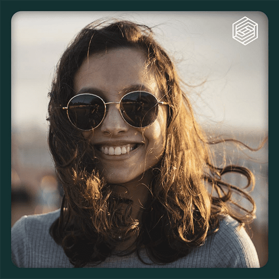 A woman with wavy brown hair smiles broadly while wearing round sunglasses. The background is blurred, suggesting an outdoor setting with sunlight casting a gentle glow on her face. The image is framed with a dark green border, and a geometric logo is in the top right corner. A woman with wavy brown hair smiles broadly while wearing round sunglasses. The background is blurred, suggesting an outdoor setting with sunlight casting a gentle glow on her face. The image is framed with a dark green border, and a geometric logo is in the top right corner.